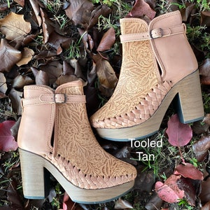 May include: A pair of tan leather ankle boots with a tooled floral design and a block heel. The boots have a buckle closure and are sitting on a bed of brown leaves.