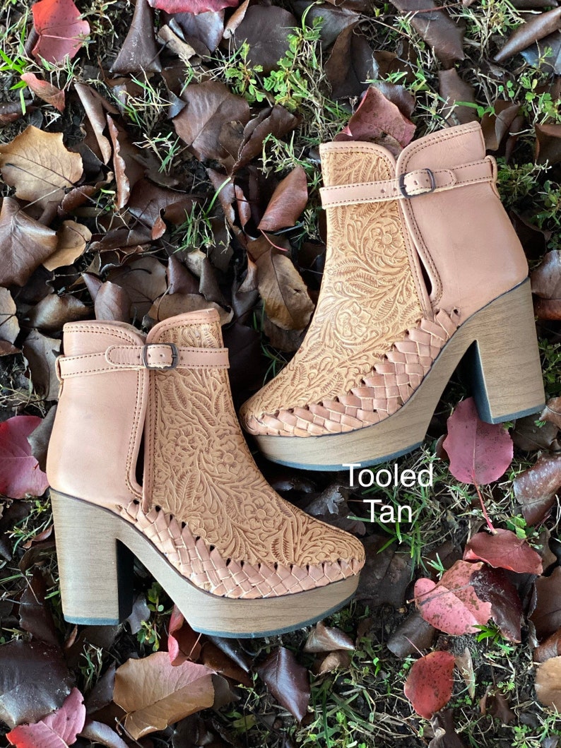 May include: A pair of tan leather ankle boots with a tooled floral design and a block heel. The boots have a buckle closure and are sitting on a bed of brown leaves.