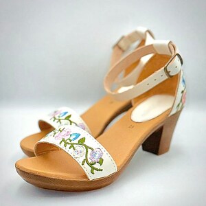 May include: White leather sandals with a floral embroidery design and a wooden heel. The sandals have an ankle strap with a buckle closure.