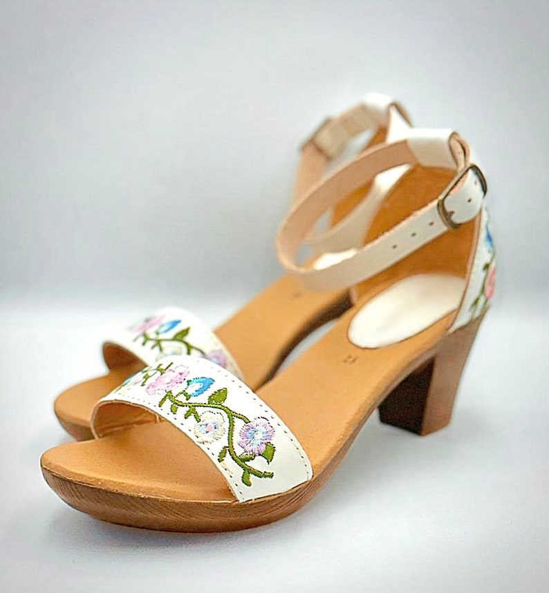 May include: White leather sandals with a floral embroidery design and a wooden heel. The sandals have an ankle strap with a buckle closure.