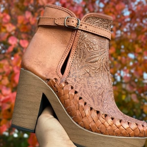 May include: Brown leather ankle boots with a woven platform sole and intricate floral embossed detailing. The boots have a buckle closure and a chunky heel.