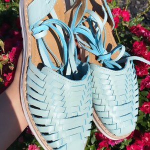 Op de afbeelding: Een paar lichtblauwe leren sandalen met geweven banden en witte zolen. De sandalen zijn gebonden met veters en hebben een kwastdetail.