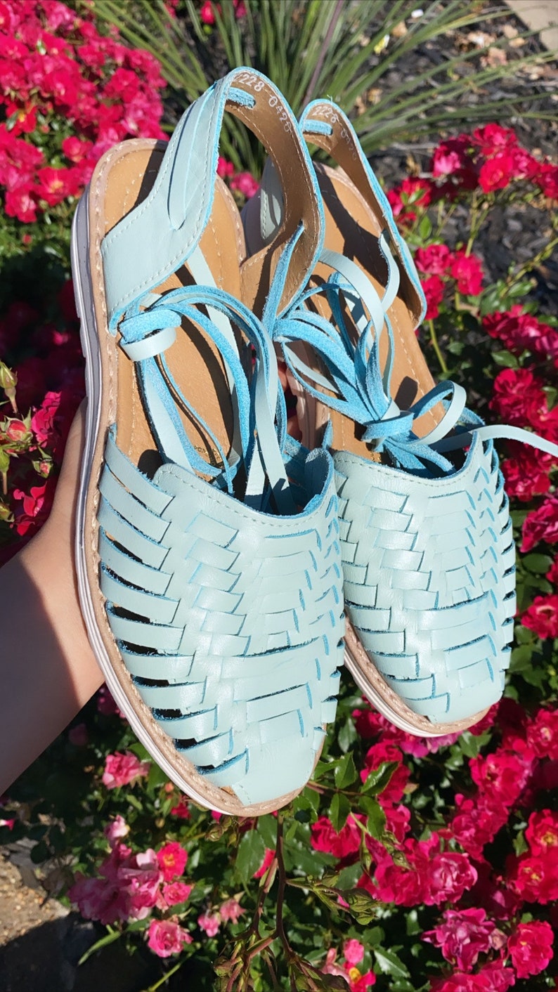 Op de afbeelding: Een paar lichtblauwe leren sandalen met geweven banden en witte zolen. De sandalen zijn gebonden met veters en hebben een kwastdetail.