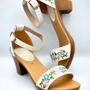 May include: White leather sandals with a wooden heel and floral embroidery. The embroidery features pink, blue, and green flowers.