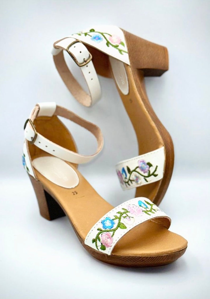 May include: White leather sandals with a wooden heel and floral embroidery. The embroidery features pink, blue, and green flowers.