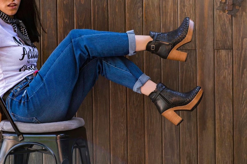 May include: A woman wearing blue jeans and black ankle boots with wooden heels sits on a metal stool. The background is a wooden wall.