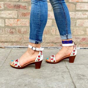 May include: White leather sandals with red floral embroidery and a low block heel. The sandals have an ankle strap with a buckle closure.