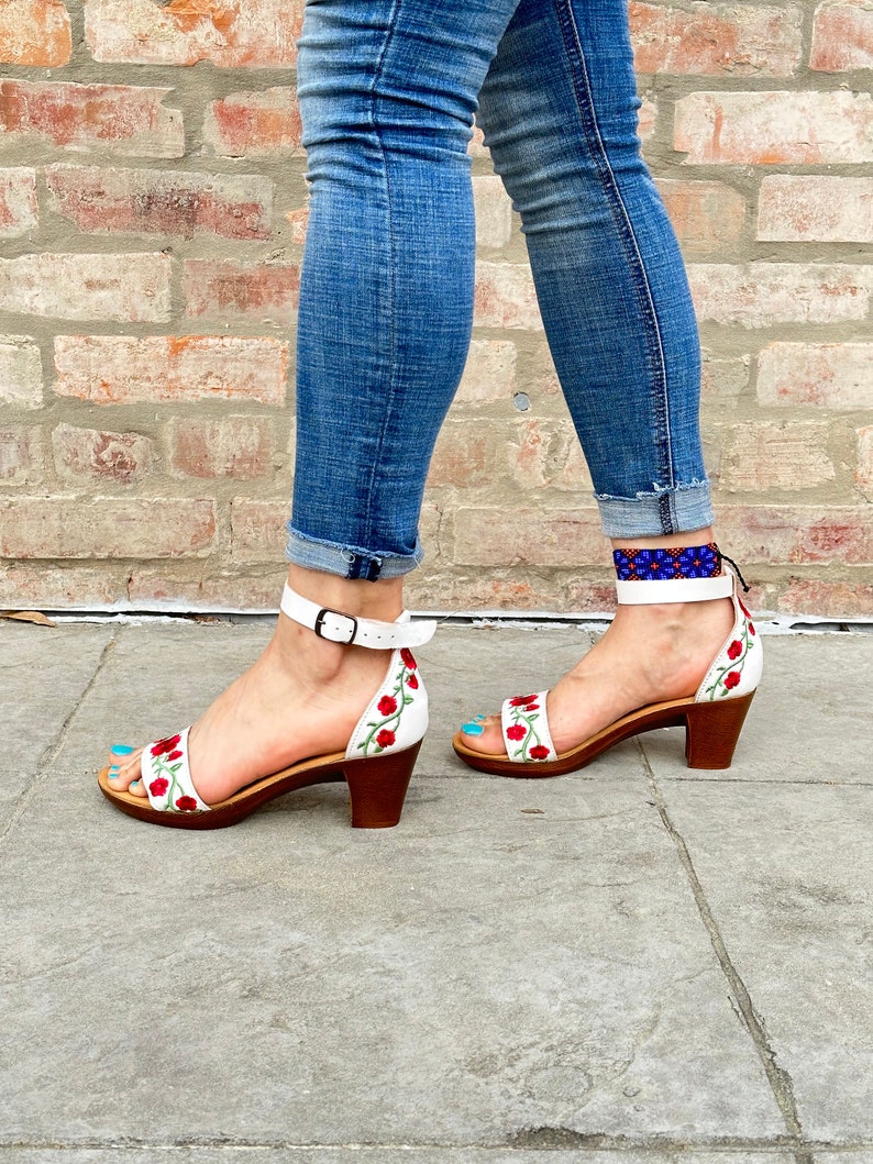May include: White leather sandals with red floral embroidery and a low block heel. The sandals have an ankle strap with a buckle closure.