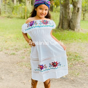 May include: A white off-the-shoulder dress with colourful floral embroidery and lace trim. The dress is worn by a young girl with brown hair and a blue and purple headband. The text "Artesanias Camila" is visible on the dress.