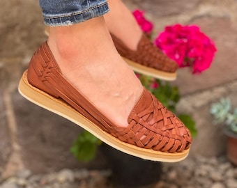 May include: Brown leather sandals with a woven design. The sandals have a flat sole and are worn on a person's feet.