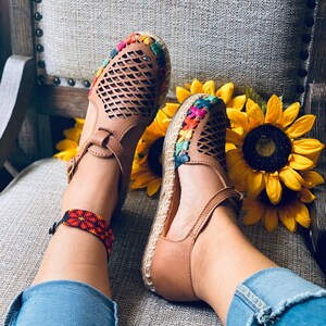 May include: A pair of brown leather sandals with colorful embroidery around the ankle strap. The sandals have a woven design and are worn by a person with a beaded ankle bracelet.