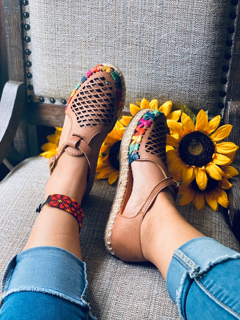 May include: A pair of brown leather sandals with colorful embroidery around the ankle strap. The sandals have a woven design and are worn by a person with a beaded ankle bracelet.