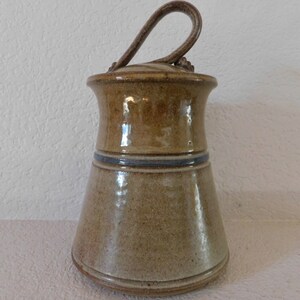 May include: A brown ceramic jar with a lid and a handle. The jar has a blue stripe around the middle.