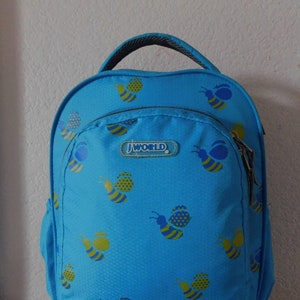May include: A blue backpack with a J World logo and a bee pattern. The backpack has a zippered pocket on the front and a handle on the top.