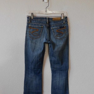 May include: A pair of blue denim jeans with a bootcut leg. The jeans have a brown leather patch on the back waistband with the text "Seven for All Mankind" in white. The back pockets have a distinctive orange stitching design.