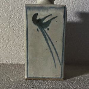 May include: A white ceramic vase with a blue bird design. The vase has a square shape and a simple, elegant design.