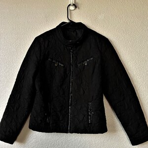May include: Black quilted jacket with a zipper closure and a stand-up collar. The jacket has two pockets with zipper closures and a decorative strap on the front.