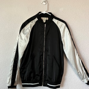 May include: Black satin bomber jacket with white satin sleeves and white trim. The jacket has a zipper closure and a ribbed collar, cuffs, and hem.