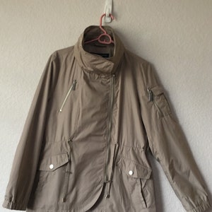 May include: A beige lightweight jacket with a zipper closure and a high collar. The jacket has two zippered pockets on the front and one zippered pocket on the left sleeve.