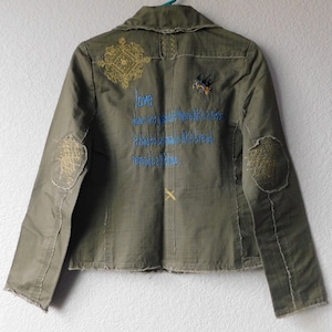 May include: A green denim jacket with embroidered details. The back of the jacket features a quote that reads "Love does not just sit there like a stone, it has to be made like bread, remade all the time."