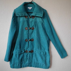 May include: Teal corduroy jacket with a ribbed knit collar and sleeves. The jacket features four brown toggle closures and two front pockets. The jacket is hanging on a black hanger.