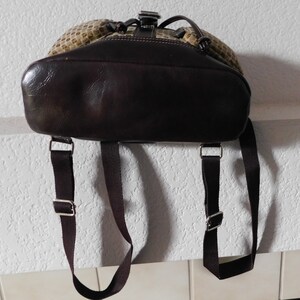 May include: A brown leather backpack with adjustable straps. The backpack has a beige and brown patterned front and a brown leather flap with a buckle closure.