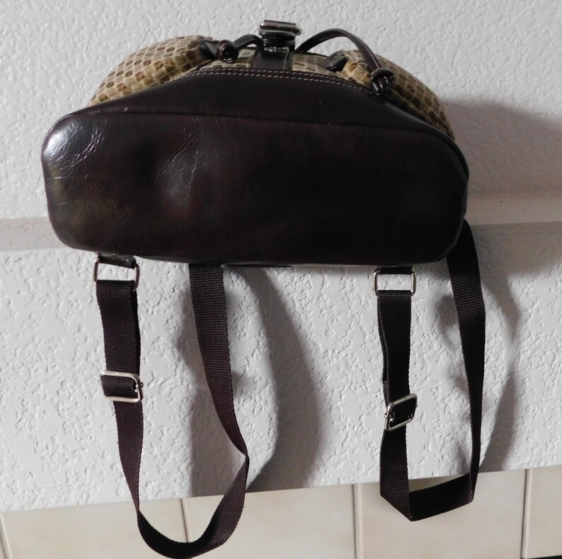 May include: A brown leather backpack with adjustable straps. The backpack has a beige and brown patterned front and a brown leather flap with a buckle closure.