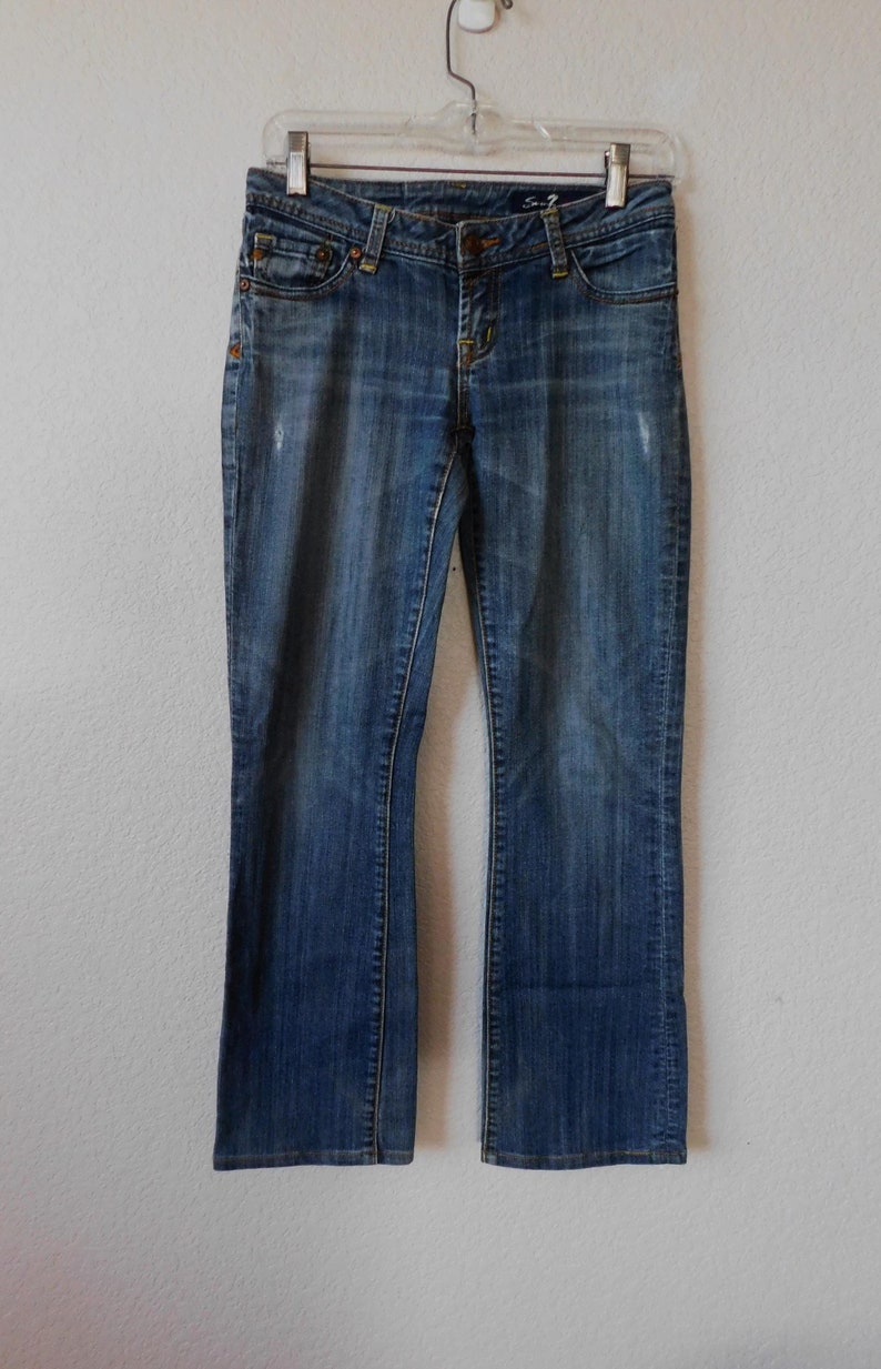 May include: A pair of blue denim jeans with a bootcut leg. The jeans have a faded look and are hanging on a clear plastic hanger.