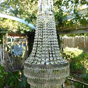 May include: A large, crystal chandelier with a silver finish hangs from a ceiling in a garden setting. The chandelier has multiple tiers of crystal drops and a round, silver base.