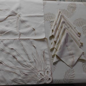 Pu&ograve; includere: Una tavola apparecchiata color crema con un sottopiatto, un tovagliolo e una pila di tovaglioli piegati. Il sottopiatto e i tovaglioli sono ricamati con un motivo floreale marrone. La tovaglia ha un motivo a foglie ripetuto.