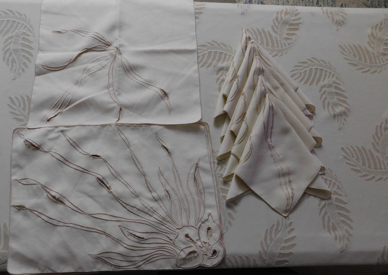 Pu&ograve; includere: Una tavola apparecchiata color crema con un sottopiatto, un tovagliolo e una pila di tovaglioli piegati. Il sottopiatto e i tovaglioli sono ricamati con un motivo floreale marrone. La tovaglia ha un motivo a foglie ripetuto.