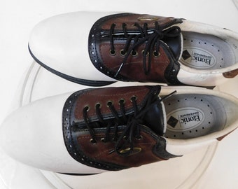 May include: Pair of white and brown leather golf shoes with black laces. The shoes have a saddle design and the brand name "Etonic" is visible on the inside of the shoes.