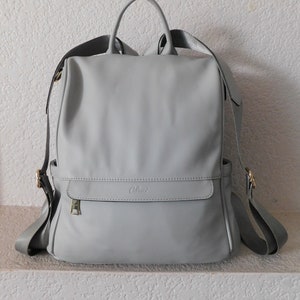 May include: A light gray leather backpack with a gold zipper and the word "Clint" embossed on the front.