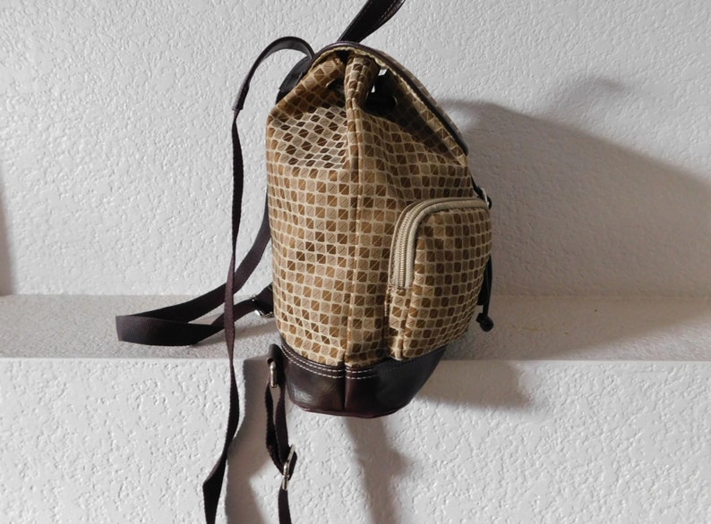 May include: A brown and beige patterned backpack with a brown leather bottom and a zippered pocket on the side.