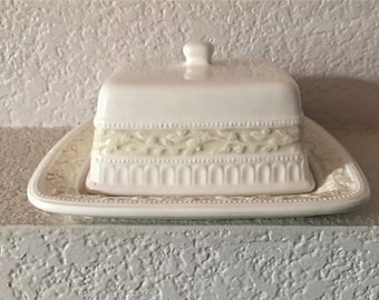 May include: White ceramic butter dish with a lid and a matching tray. The dish has a raised, decorative border with a floral design.