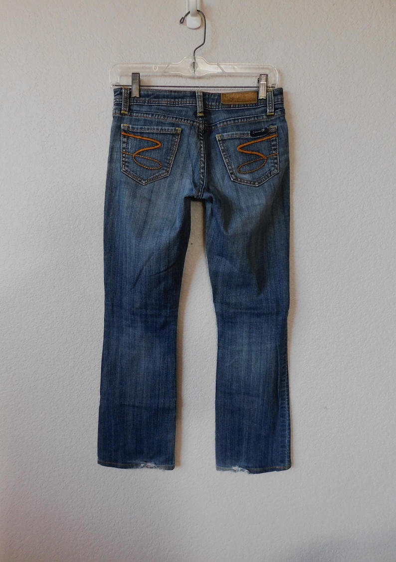 May include: A pair of blue denim jeans with a bootcut leg. The jeans have a brown leather patch on the back waistband with the text "Seven for All Mankind" in white. The back pockets have a distinctive orange stitching design.