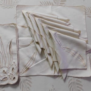 Pu&ograve; includere: Un set di tovaglioli e tovagliette in lino color avorio. Le tovagliette presentano un design floreale ricamato con dettagli marroni. I tovaglioli sono piegati e impilati, mostrando l'intricato ricamo. Ideale per cene formali.