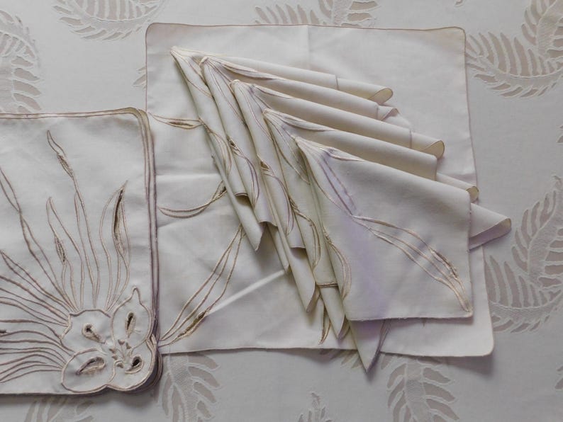 Pu&ograve; includere: Un set di tovaglioli e tovagliette in lino color avorio. Le tovagliette presentano un design floreale ricamato con dettagli marroni. I tovaglioli sono piegati e impilati, mostrando l'intricato ricamo. Ideale per cene formali.