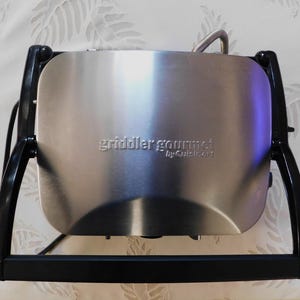 May include: A stainless steel griddler gourmet by Cuisinart. The appliance has a rectangular cooking surface and a black frame. The text "griddler gourmet" is embossed on the front. The griddler has a handle and a power cord.