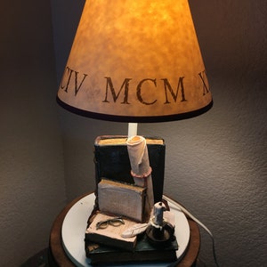 May include: A lamp with a brown lampshade that says "CIV MCM X" on it. The base of the lamp is a stack of books with a quill pen, glasses, and a candle on top.
