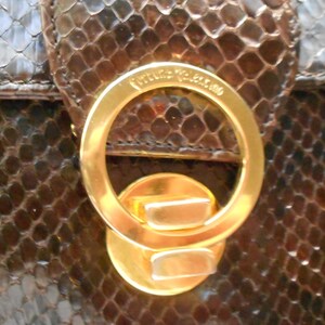 May include: Close-up of a gold-toned metal clasp with the text "Valentino" engraved on the top. The clasp is attached to a brown snakeskin handbag.