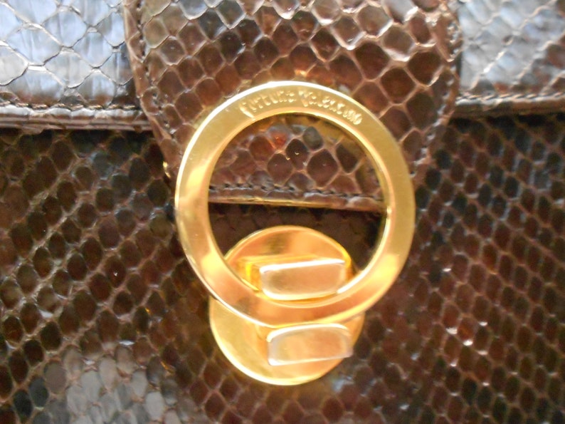 May include: Close-up of a gold-toned metal clasp with the text "Valentino" engraved on the top. The clasp is attached to a brown snakeskin handbag.