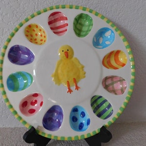 May include: A white ceramic Easter egg plate with a yellow chick in the center. The plate has 12 egg-shaped indentations around the edge, each decorated with a different colorful pattern.