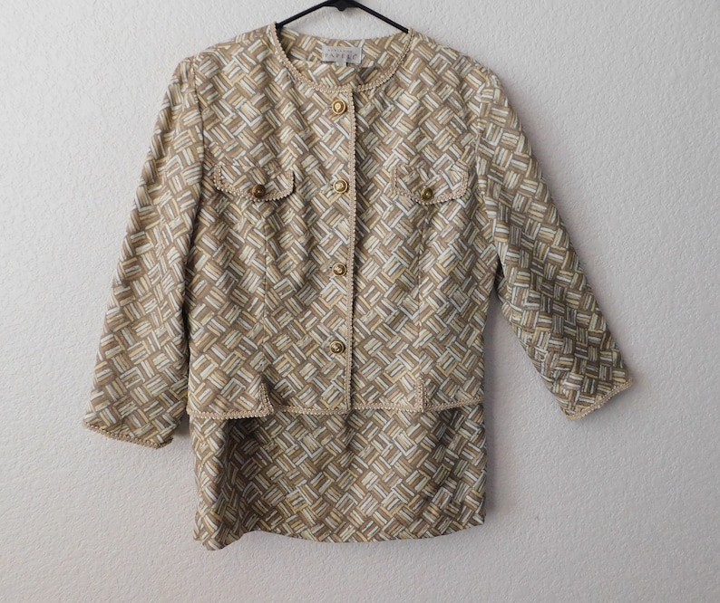 May include: A beige and brown patterned jacket with gold buttons and a matching skirt. The jacket has a round neckline and three-quarter sleeves. The skirt is knee-length.