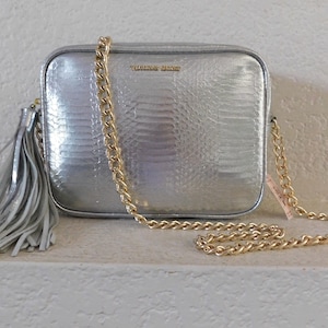 May include: A silver chain strap crossbody bag with a silver tassel. The bag has a snakeskin embossed texture and the brand name "Victoria's Secret" is printed on the front.