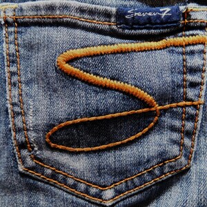 May include: Close-up of a blue denim pocket with a brown stitched design. The pocket has a small label that reads "Seven7" in white lettering.