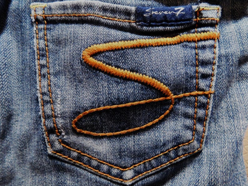 May include: Close-up of a blue denim pocket with a brown stitched design. The pocket has a small label that reads "Seven7" in white lettering.