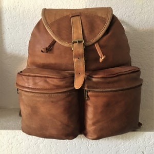 May include: A brown leather backpack with two large front pockets and a buckle closure. The backpack has a flap with a buckle closure and a drawstring.