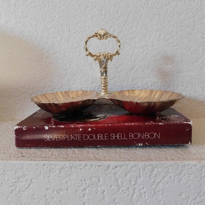 May include: A silver-plated double shell bonbon dish with a decorative handle. The dish has two shell-shaped bowls and sits on a red box. The handle is ornate and features a looped design.