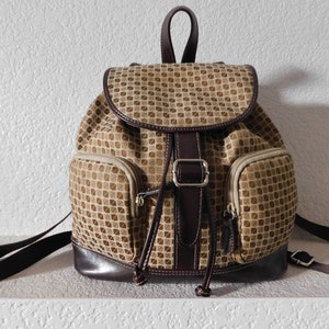 May include: A brown and tan patterned backpack with a brown leather trim and a silver buckle. The backpack has two front pockets and a drawstring closure.